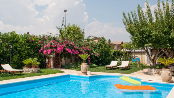 A bright blue swimming pool with floating loungers atop. Bright green foliage surrounds the pool along with bright purple flowers.