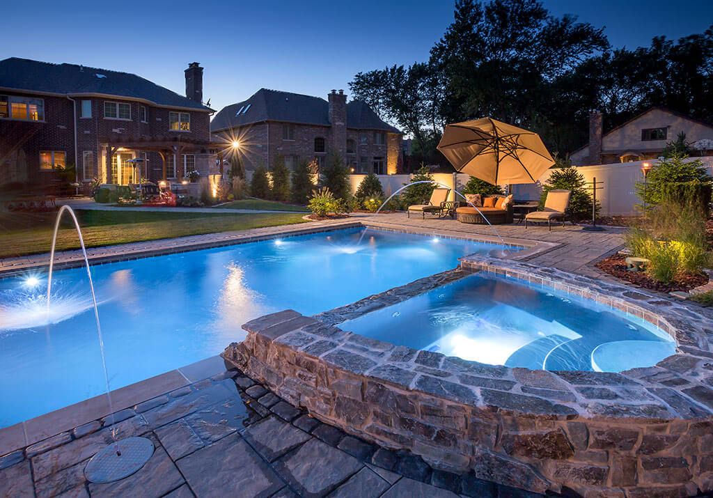 Lit up backyard pool with a fountain at dusk